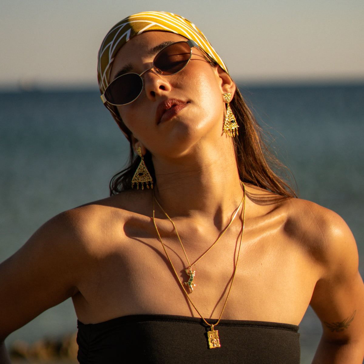 Model by the sea in a headscarf wearing the Exit engraved gold pendant with layered necklaces and gold earrings