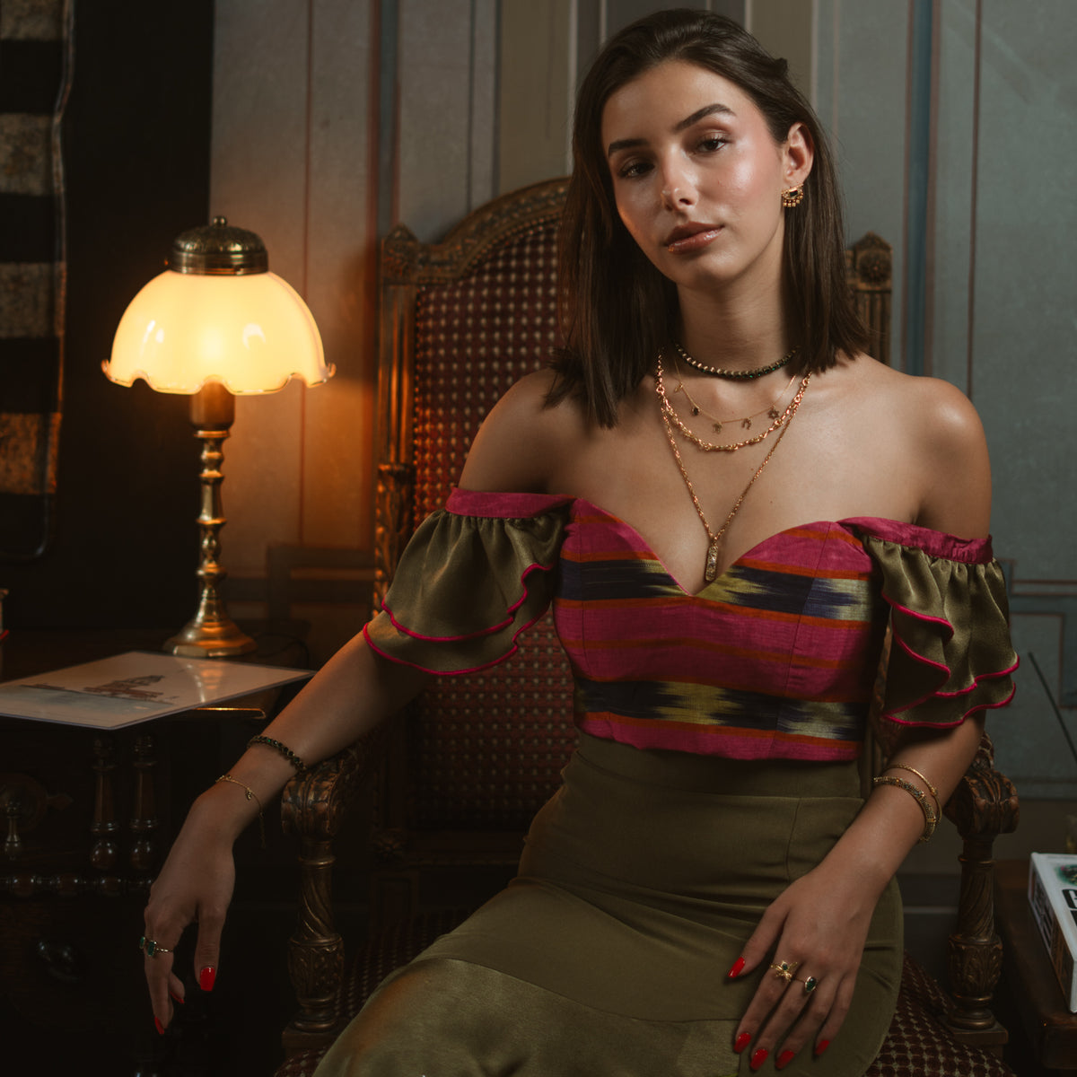 Model seated in a striped off shoulder top wearing the Five Guardians gold charm necklace in a layered look