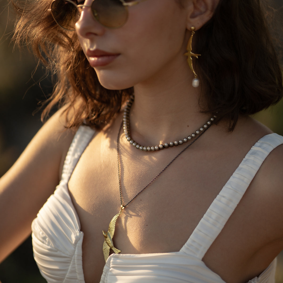 Woman in sunglasses and a white dress wearing the Free Spirit Sparrow Gold Pendant Two Tone Necklace with the matching sparrow pearl drop earrings
