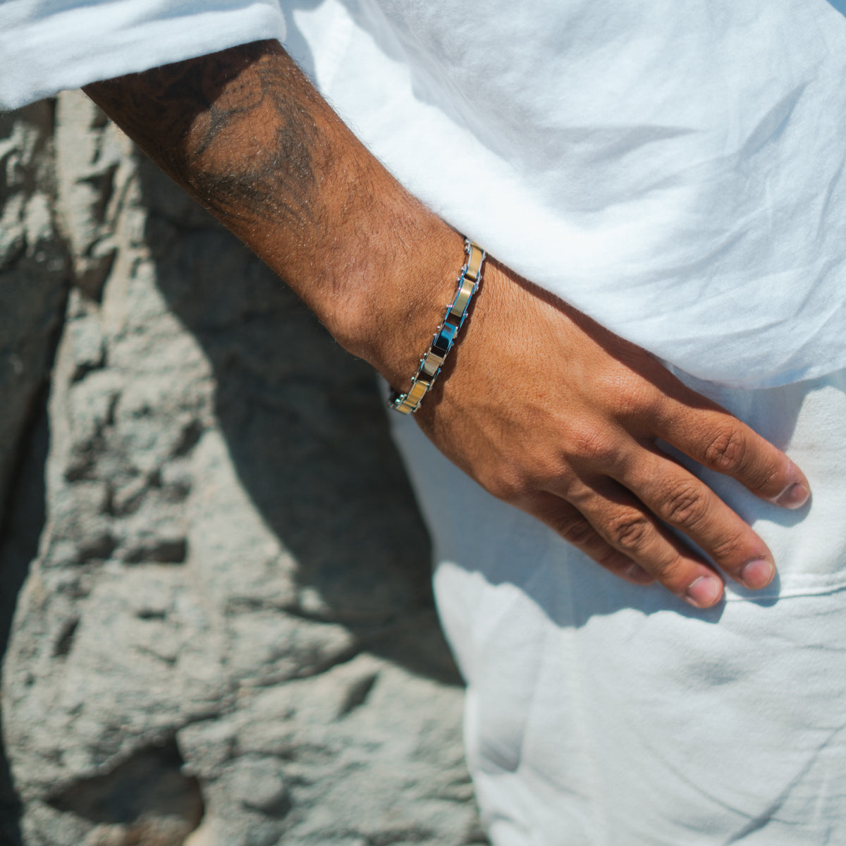Man in white clothing wearing the two-tone steel link bracelet, hand resting at his side against a stone wall