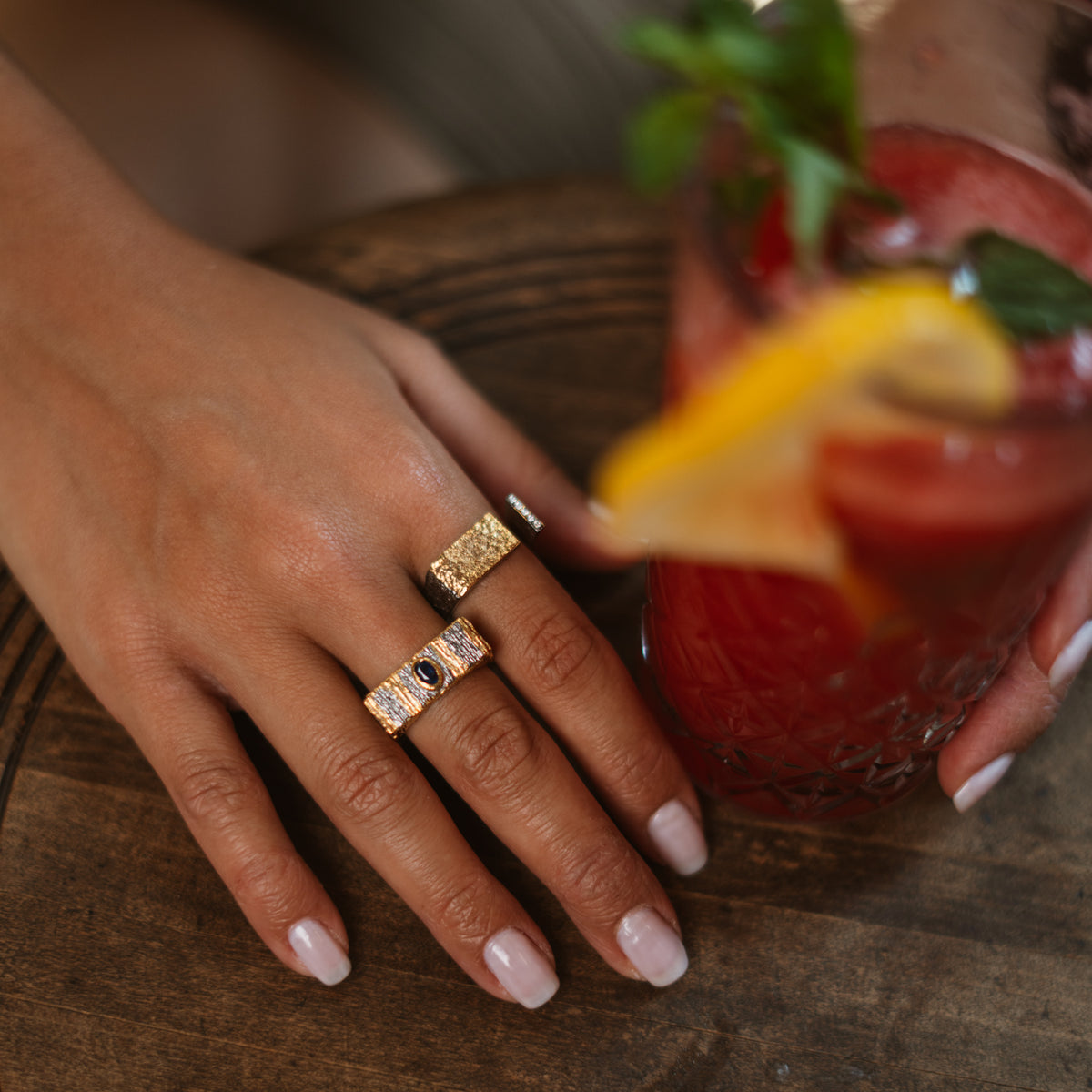 Hand on a wooden table beside a red drink, wearing the Golden Contrast ring stacked with the Midnight Temple two-tone sapphire ring