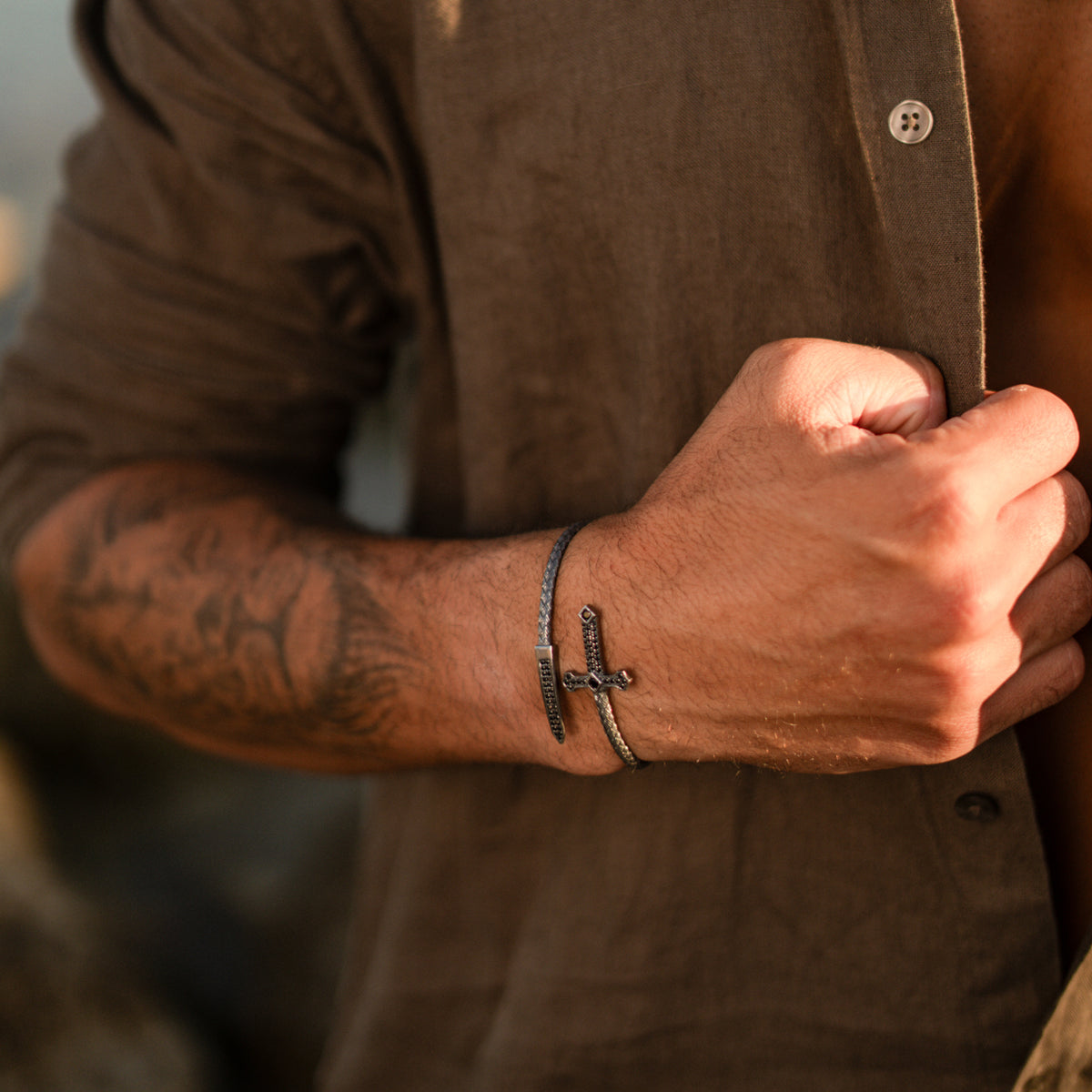 Close view of the Gothic cross and dagger silver wrap bracelet worn on a man's wrist, hand gripping a linen shirt