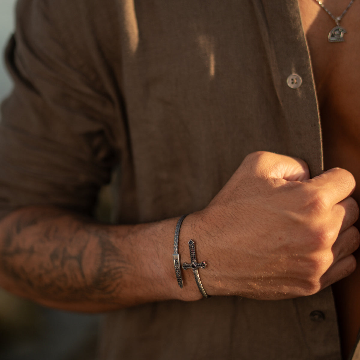 Men's cross and dagger wrap bracelet worn with an open brown linen shirt and a silver pendant necklace
