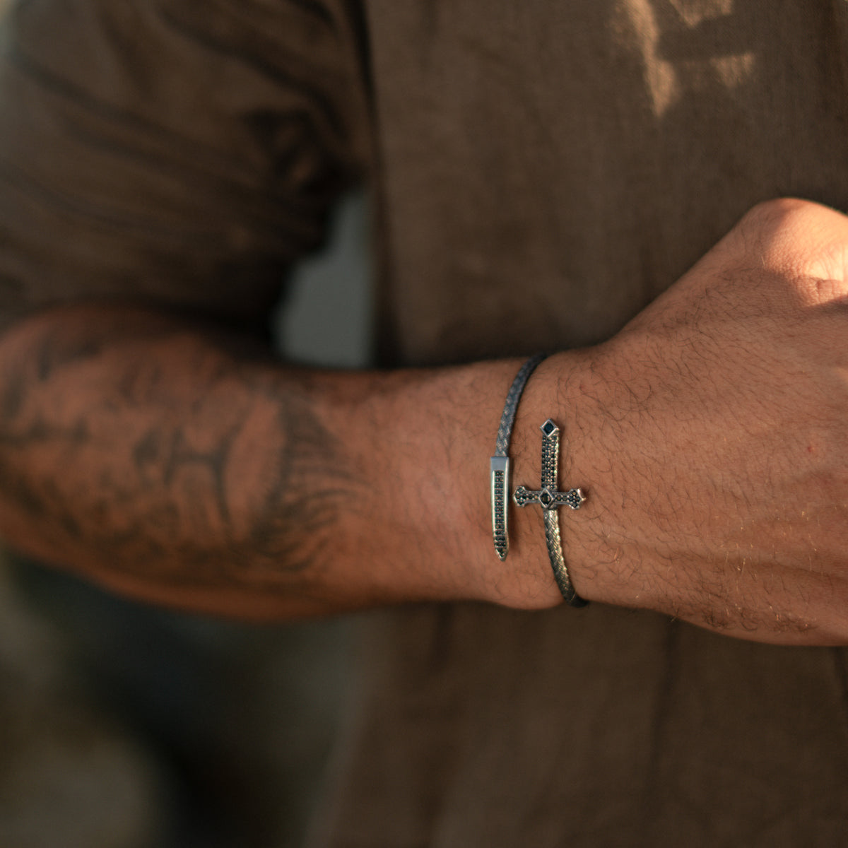 Man in a brown linen shirt wearing the oxidized sterling silver cross and dagger wrap bracelet on his tattooed forearm