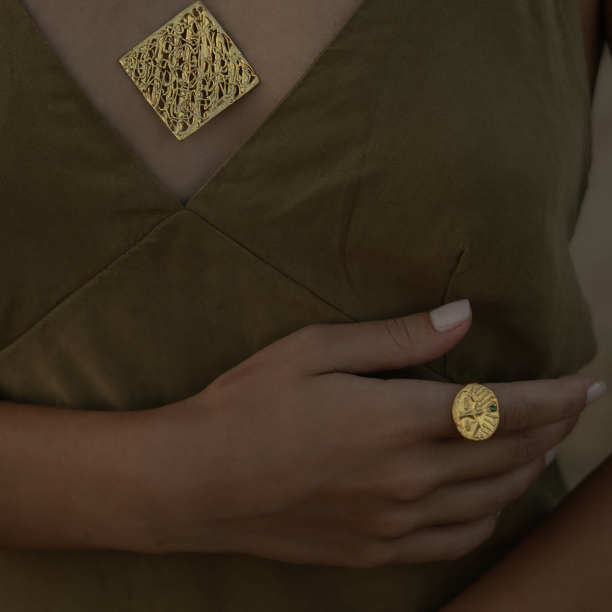 Hand on an olive dress wearing the Guardian Emerald Eye gold ring, a textured gold square pendant at the neckline