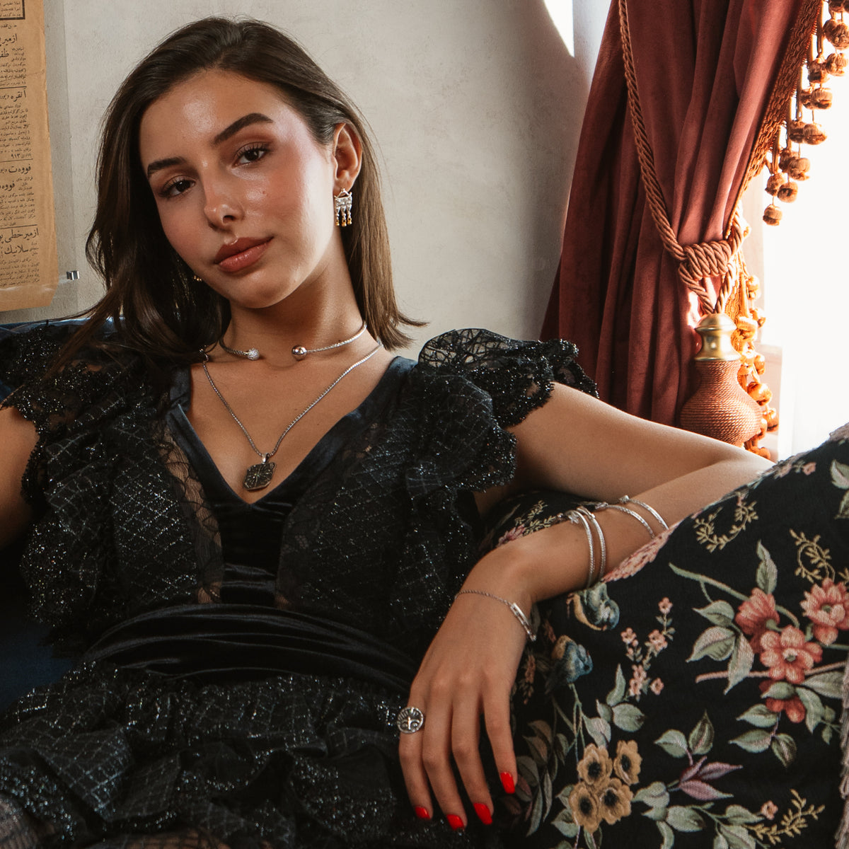 Model in a black lace dress on a floral sofa wearing the Guardian Emerald Eye sterling silver ring with layered silver necklaces