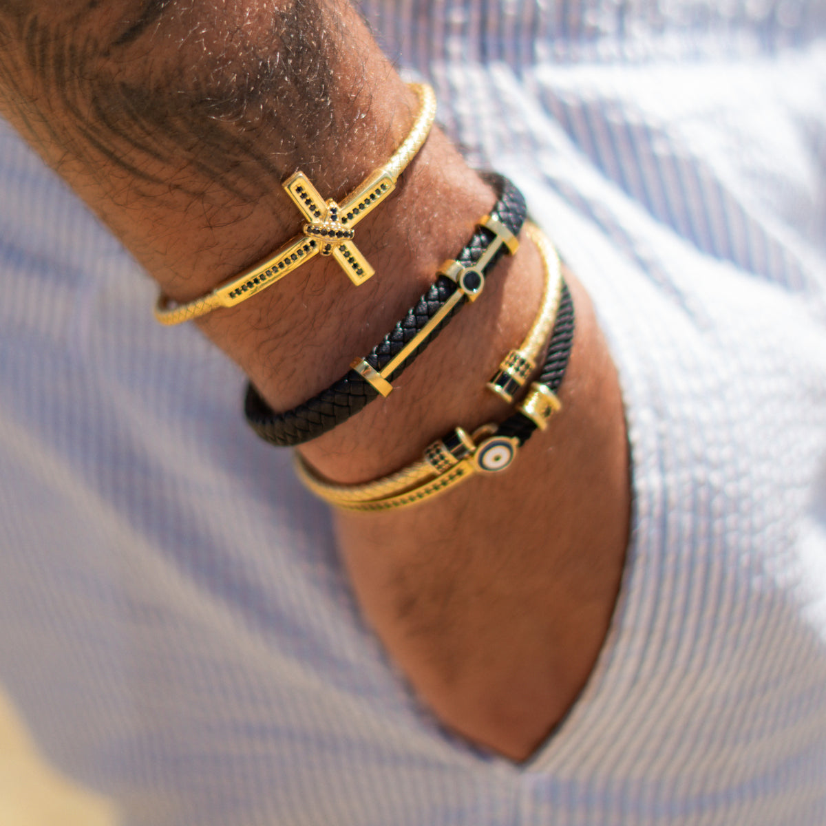 Stack of men's gold bracelets on a wrist in a white striped shirt, with the evil eye black cord bracelet at the bottom