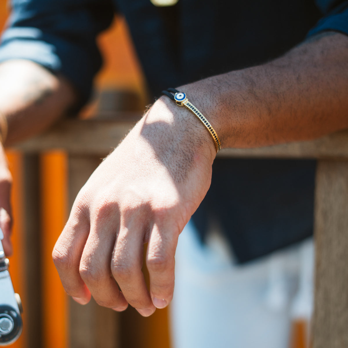 Man in a navy shirt leaning on a wooden rail, wearing the black cord gold evil eye bracelet in sunlight
