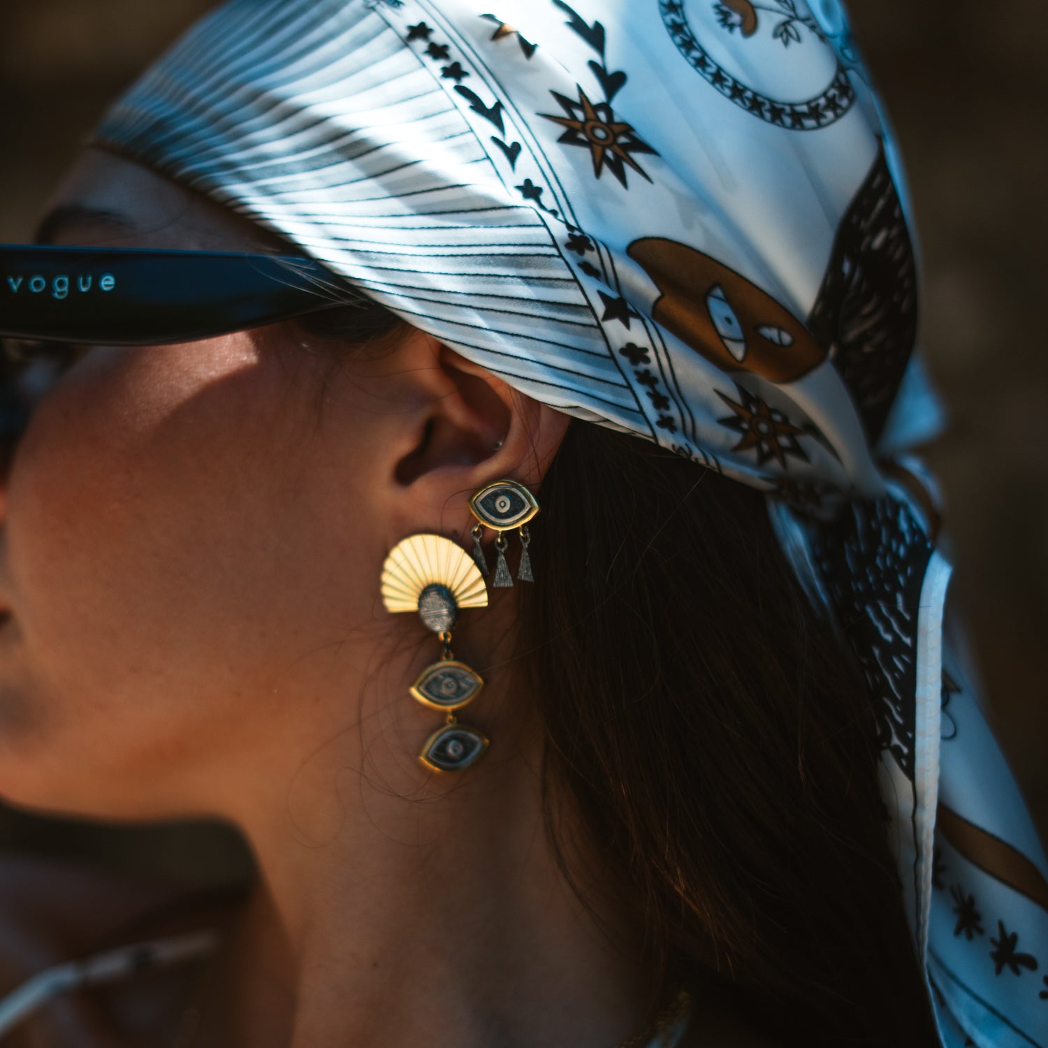 Close-up of a model's ear wearing the Guardian Eye Fringe stud earring with a headscarf and sunglasses in the background