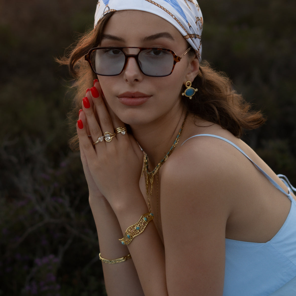 Model in a headscarf and blue dress wearing the Honeycomb Lace Turquoise & Ruby Gold Cuff Bracelet with layered gold necklaces