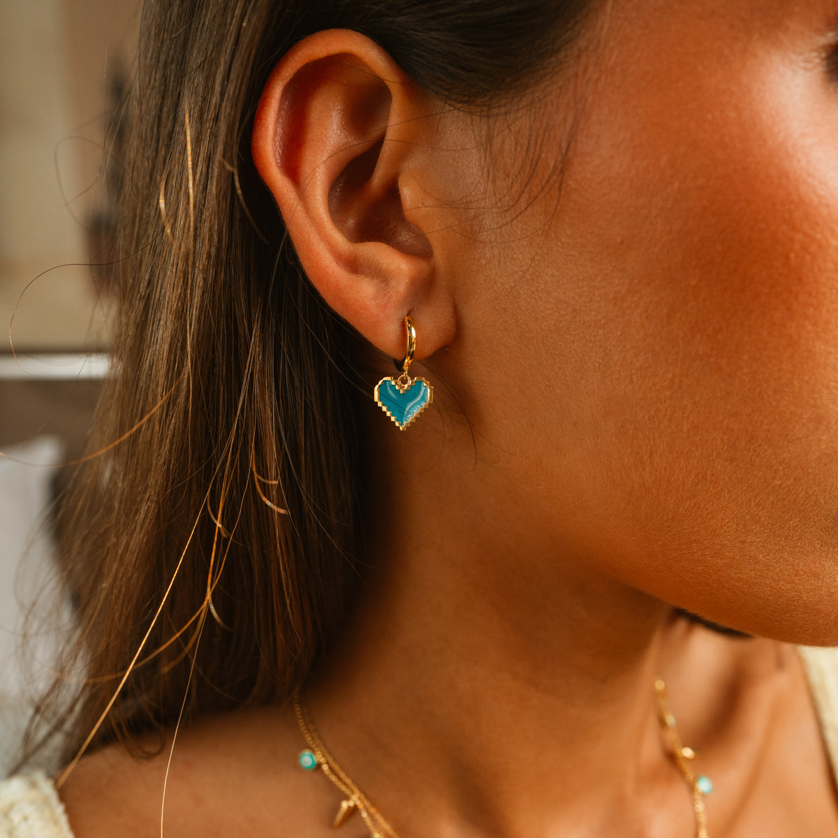 Close up of a turquoise enamel heart gold hoop earring on the ear, layered with a fine gold charm necklace