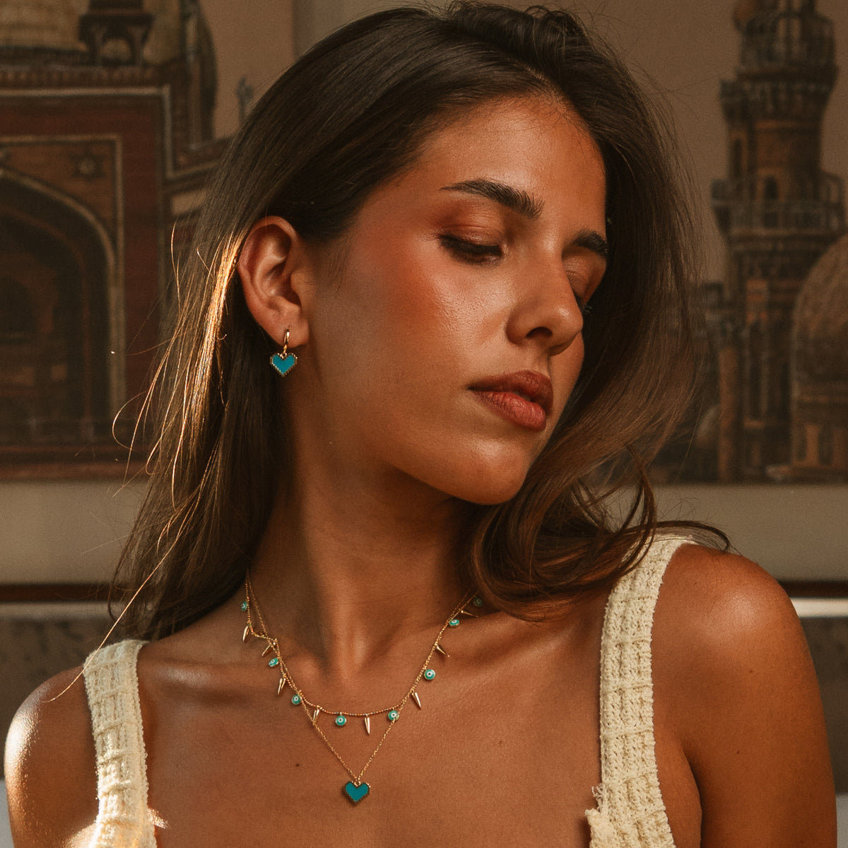 Model looking down wearing the turquoise enamel heart gold hoop earrings with a layered gold necklace look