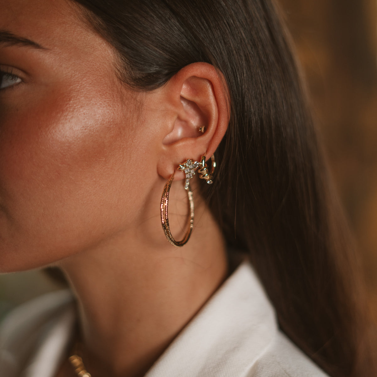 Close view of a model's ear wearing the gold love knot huggie stacked with a baguette star stud and a large twisted gold hoop