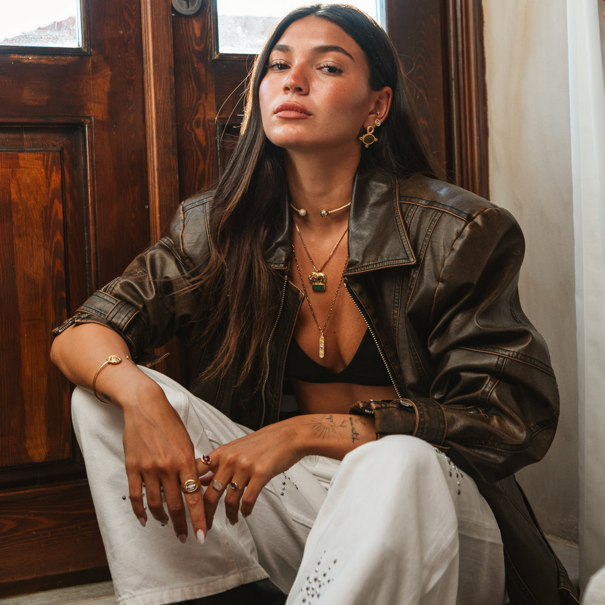 Model in a brown leather jacket seated by a wooden door wearing the green mosaic gold elephant pendant