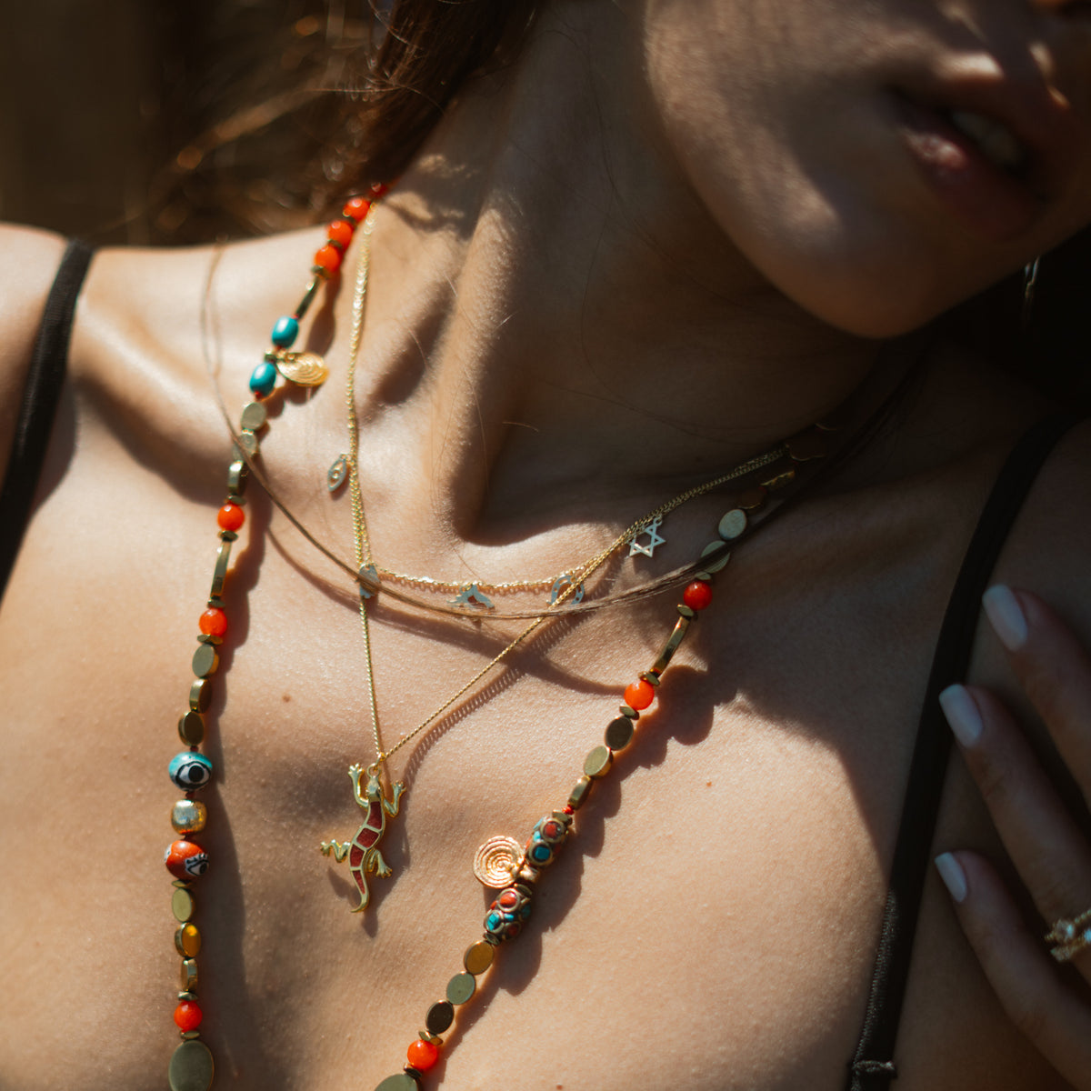 Woman in sunlight wearing the red enamel gecko pendant necklace with layered gold and beaded necklaces