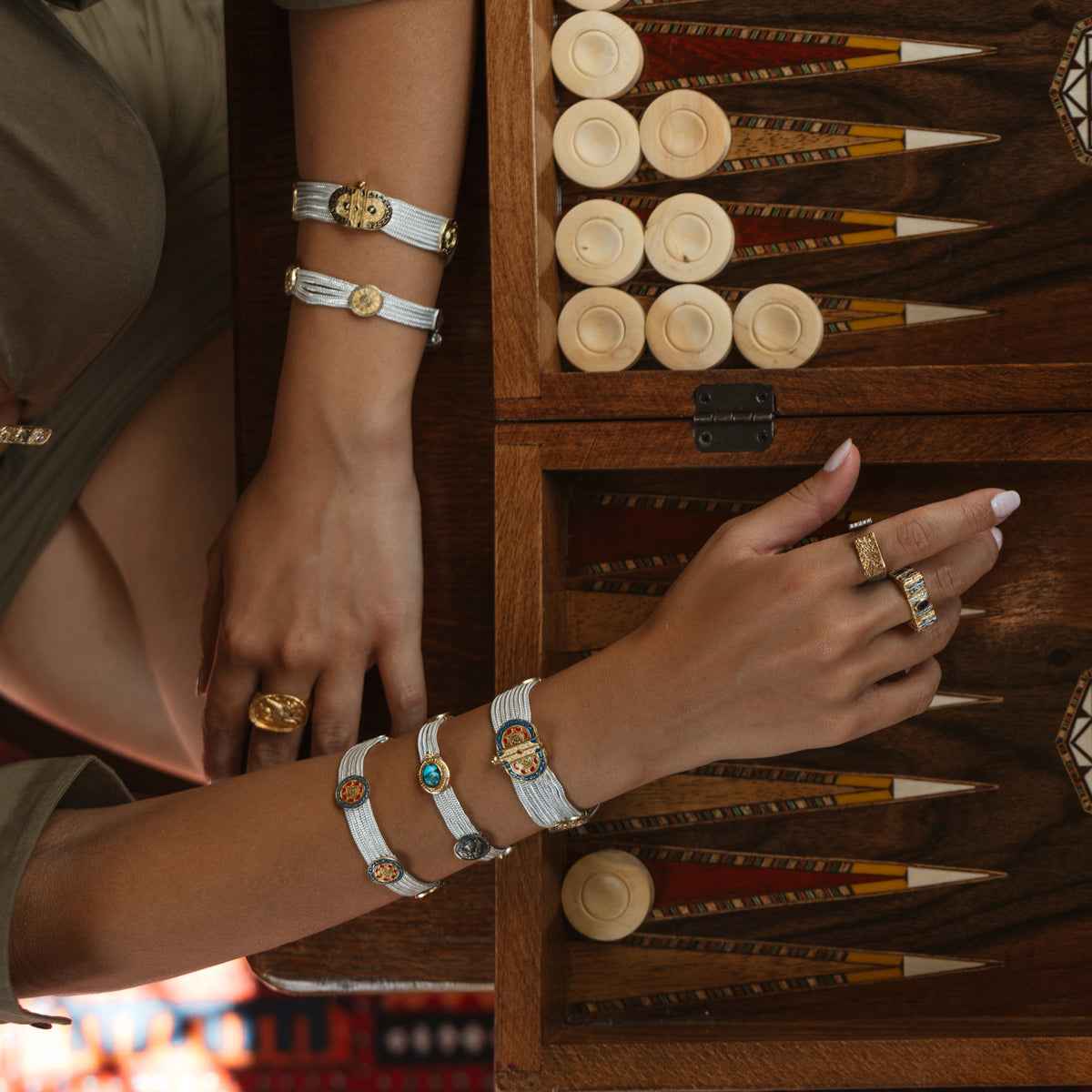 Hands over a backgammon board, the Midnight Temple two-tone ring worn with woven silver medallion bracelets