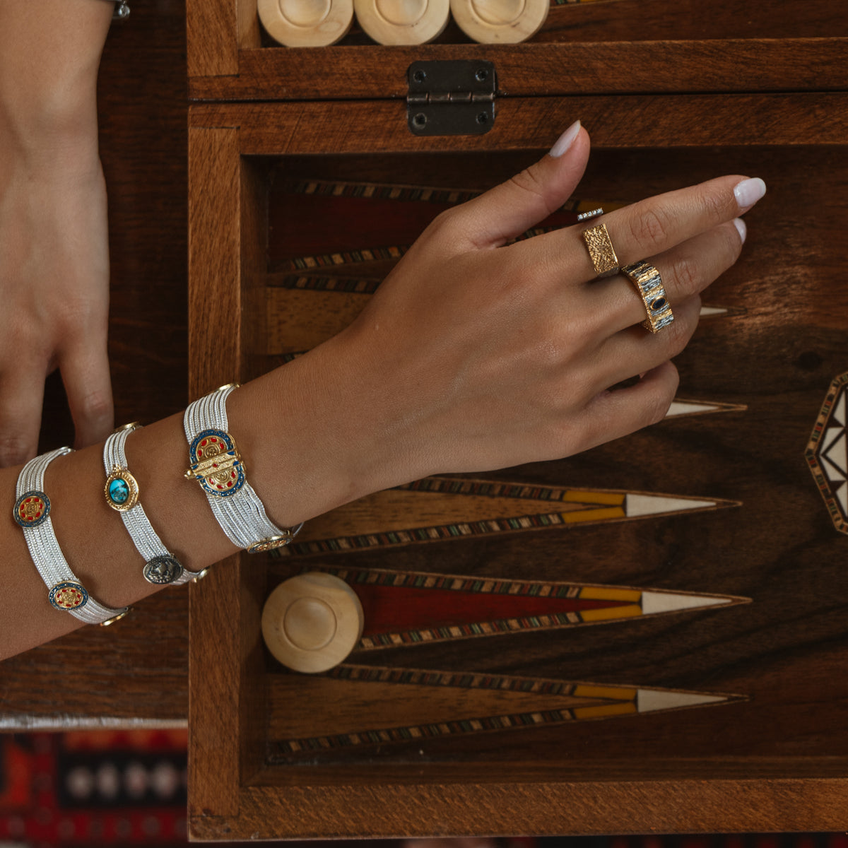Hand resting on an open backgammon board, the Midnight Temple sapphire ring stacked with a gold ring and woven bracelets