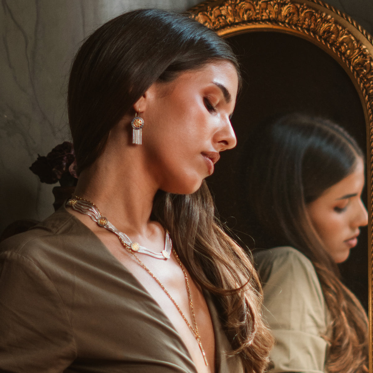 Model in profile beside a gilded mirror wearing the Mystic Bloom tassel earring with a woven silver collar necklace