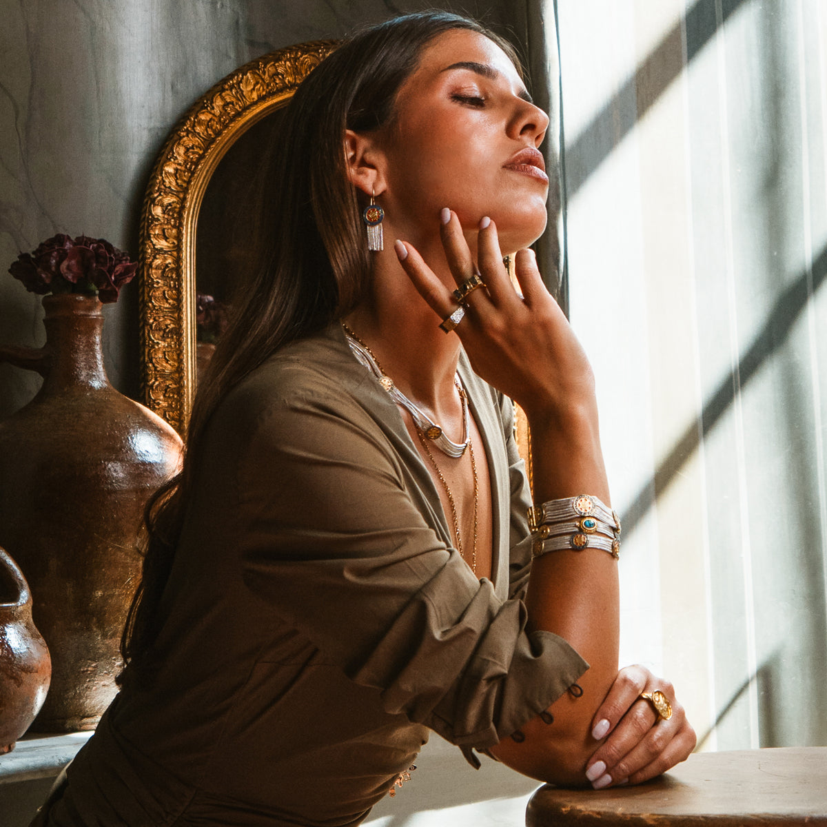 Model with her hand at her jaw by a sunlit window, wearing the Mystic Bloom tassel earrings with stacked woven two-tone bracelets