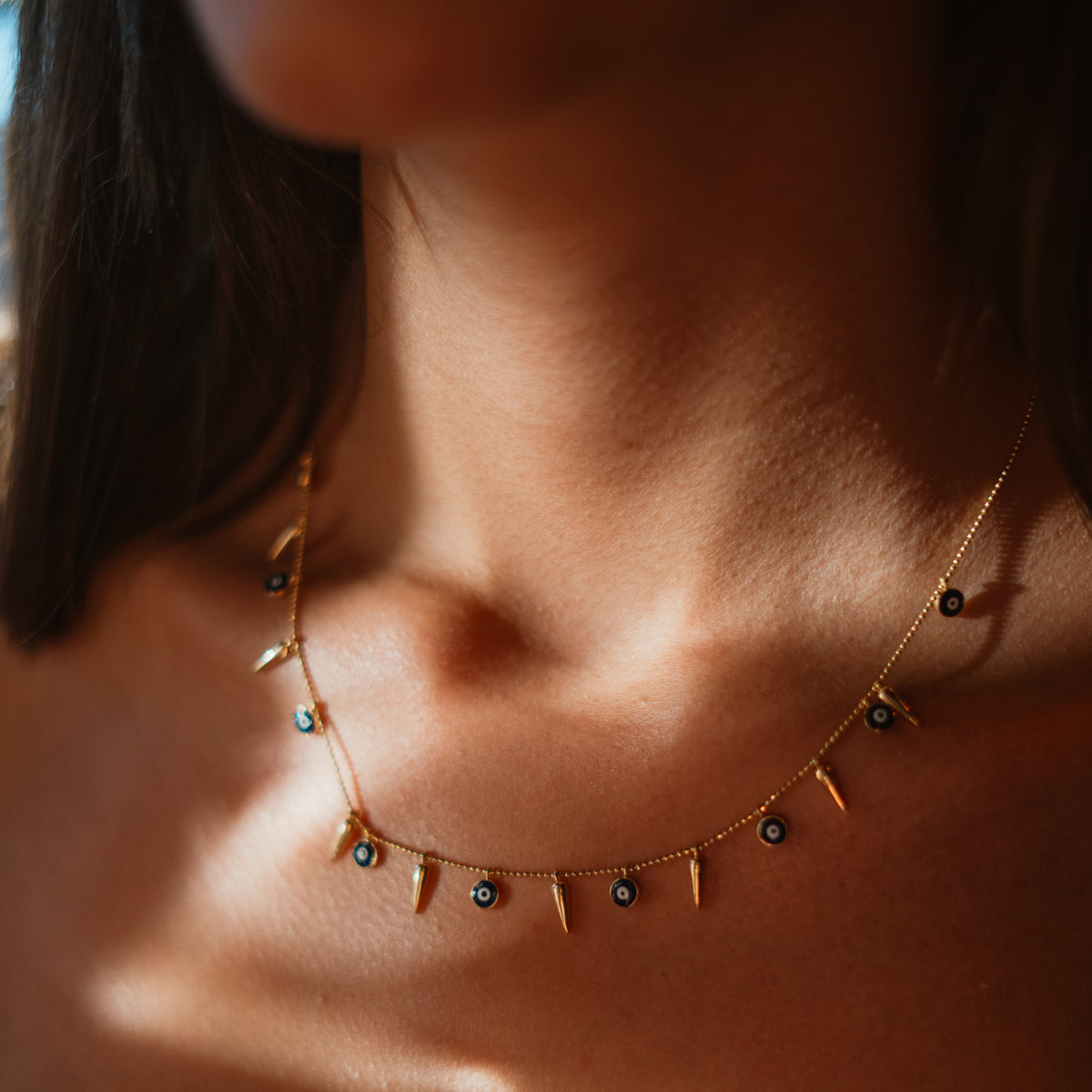 Close up of the navy evil eye gold spike charm necklace resting on the collarbone in warm light