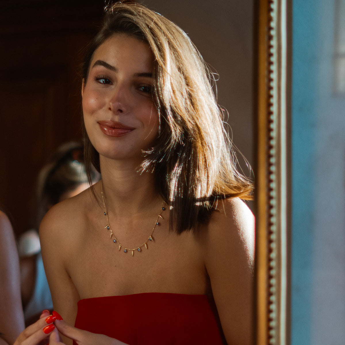 Smiling model in a red strapless dress wearing the navy blue evil eye and gold spike charm necklace