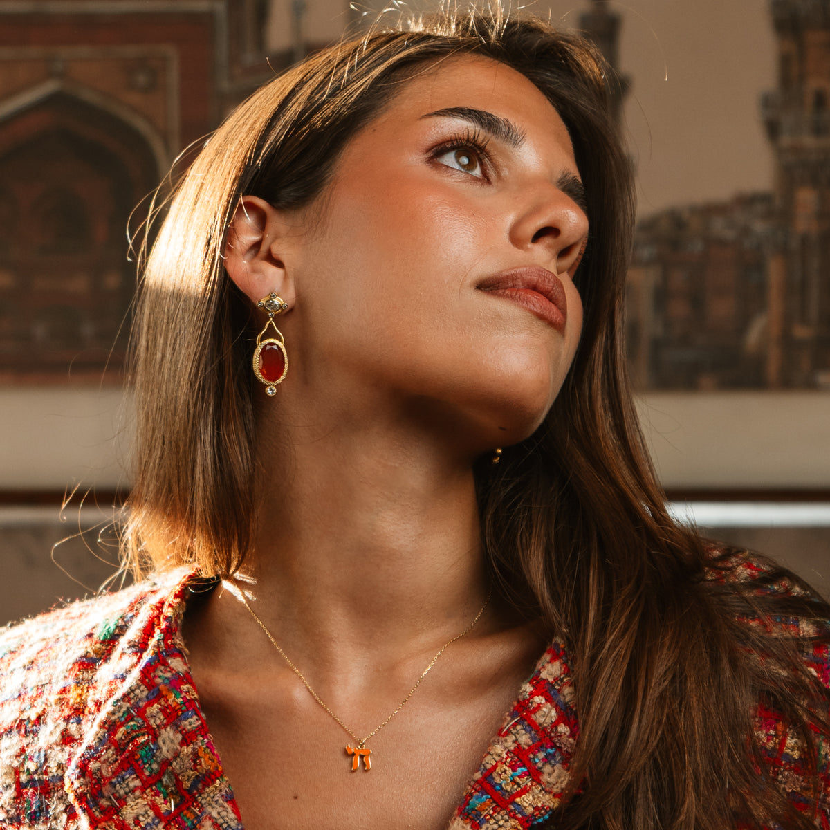 Woman looking up in warm light wearing the orange Chai gold pendant necklace and red stone drop earrings