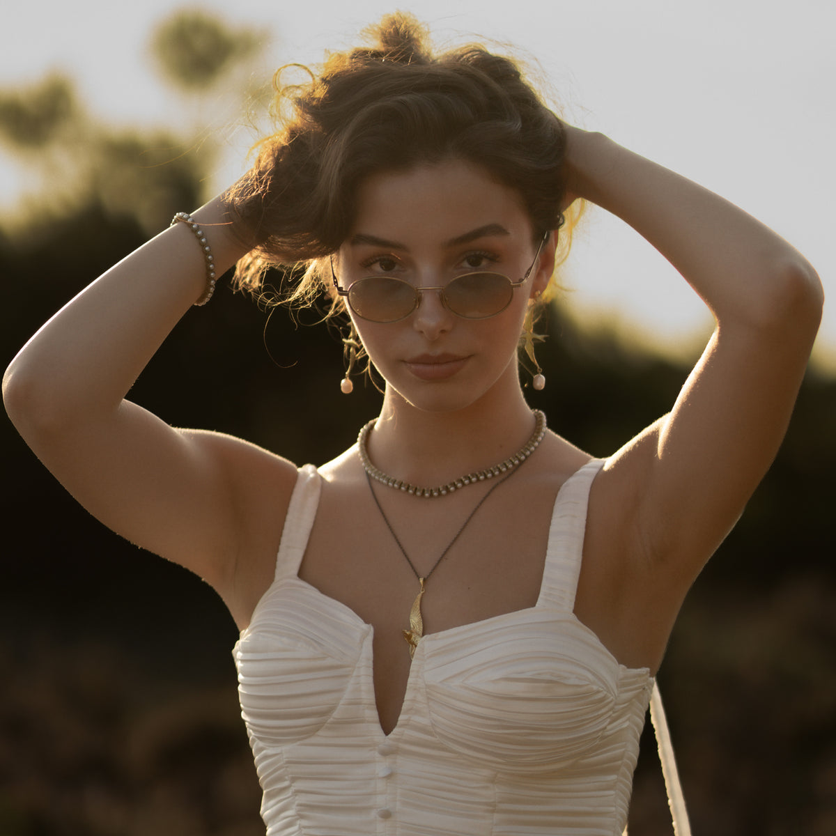 Model with arms raised wearing the antique gold pearl tennis bracelet, pearl drop earrings and a layered gold necklace