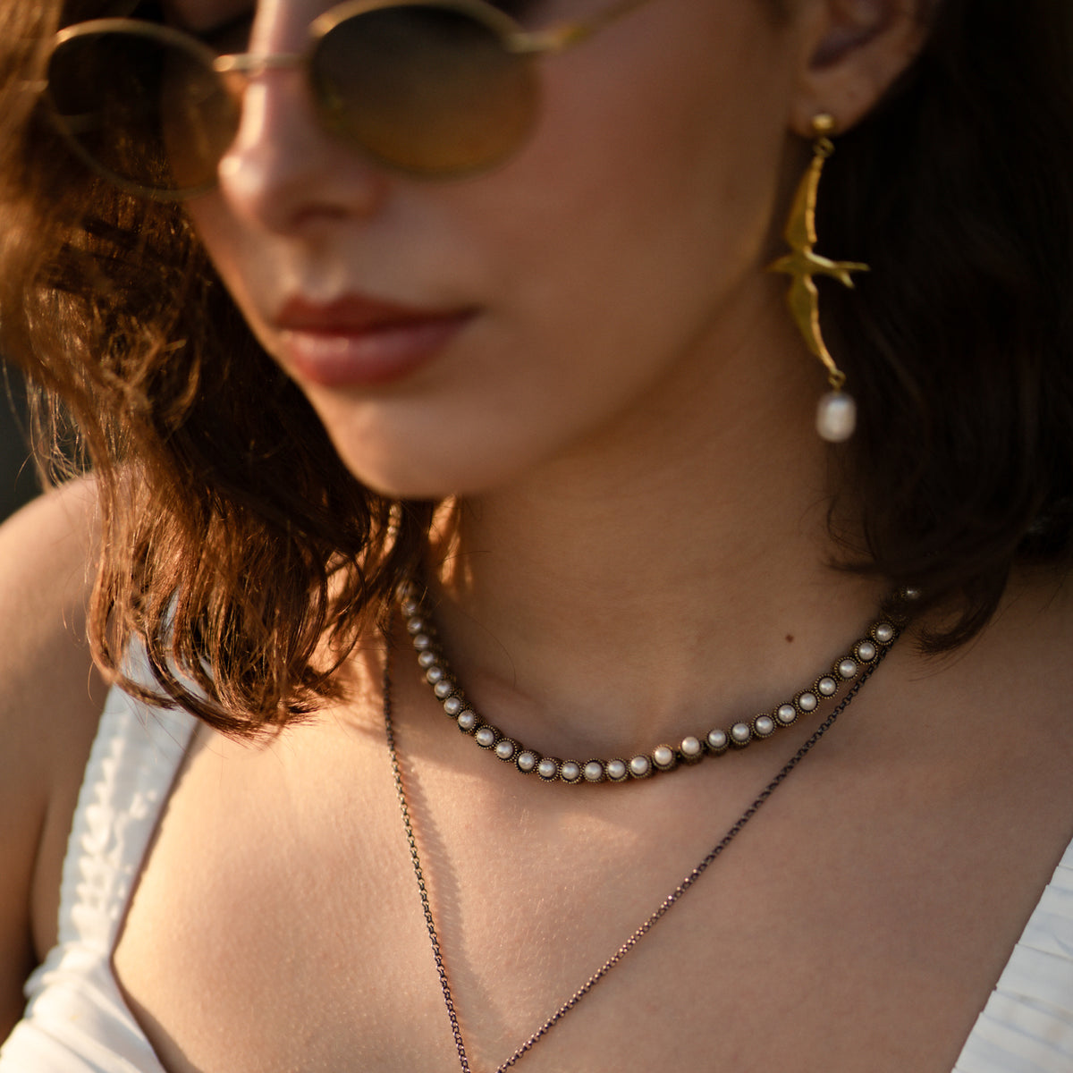 Close up of the pearl tennis necklace worn at the base of the neck at golden hour, layered over a longer gold chain