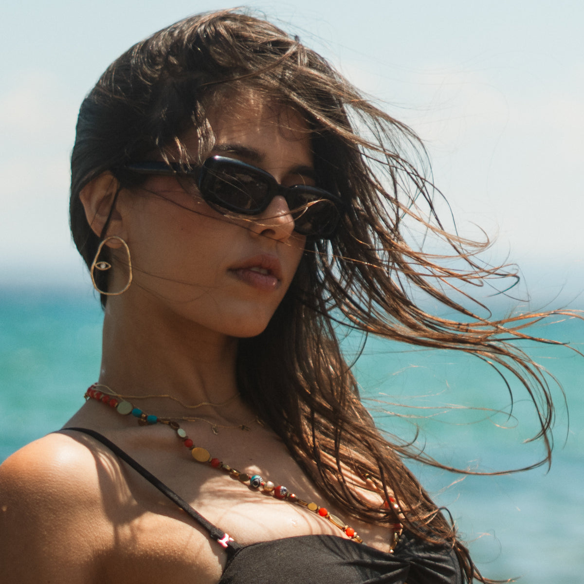 Model at the beach wearing the Protective Halo Evil Eye Gold & Diamond Hoop Earrings, the gold circle catching the light