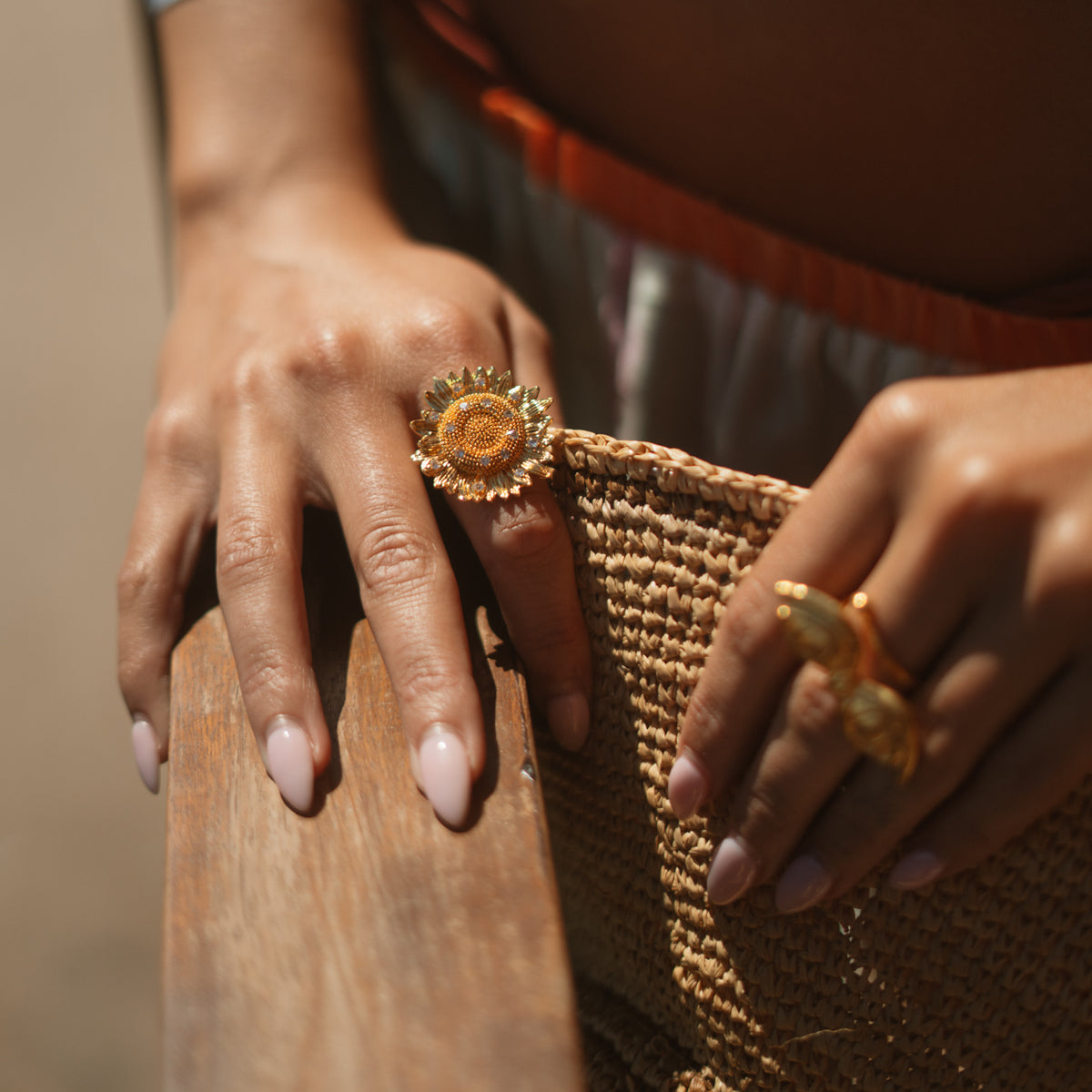 Hands resting on weathered wood, the gold sunflower ring catching sunlight next to a straw tote