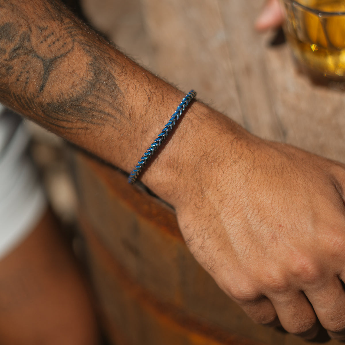 Blue cord foxtail steel bracelet on a tattooed wrist resting on a wooden bar beside a glass