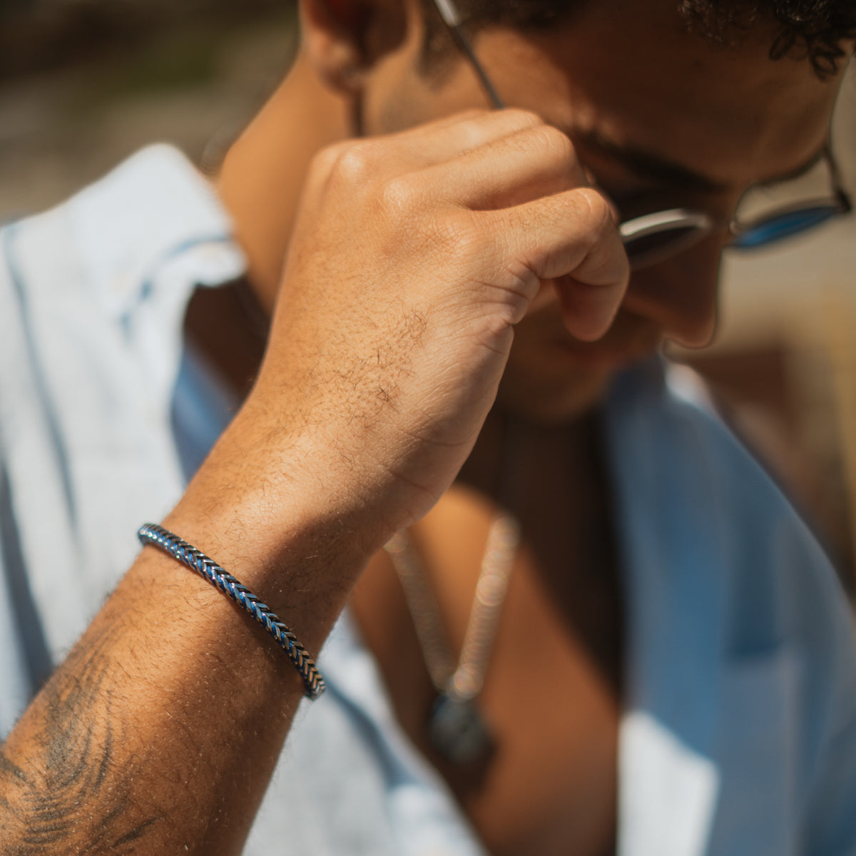 Man in sunglasses and a light blue shirt wearing the blue cord foxtail steel bracelet, hand raised to his face