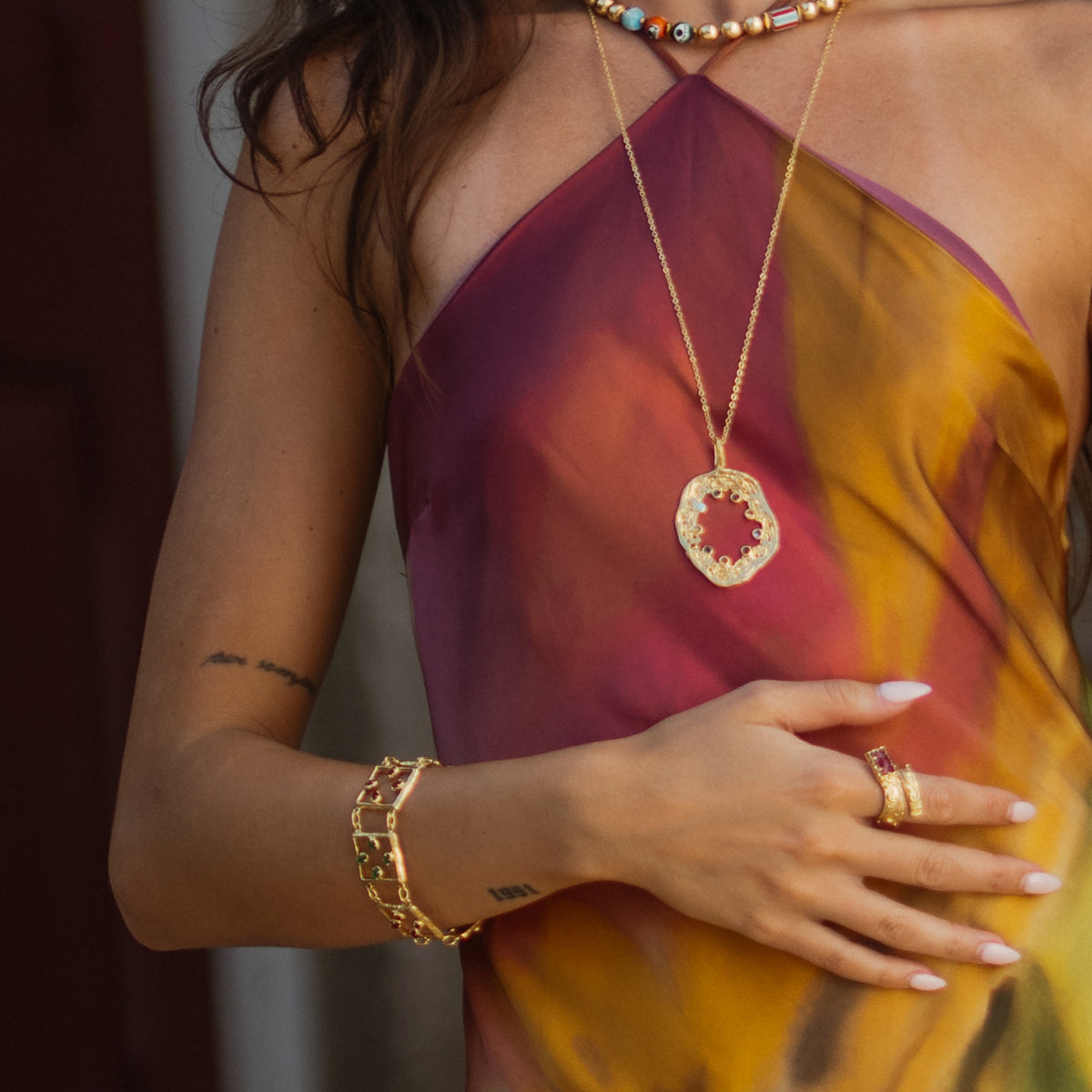Model in a sunset toned halter dress wearing the gemstone gold link bracelet with a gold pendant necklace and stacked rings