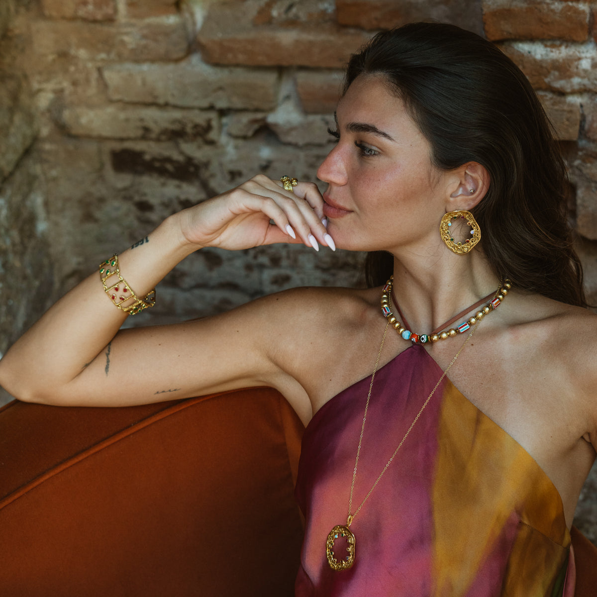 Model against a brick wall wearing the Ruby & Emerald Gemstone Open Square Gold Link Bracelet with gold hoop earrings and a gold pendant necklace