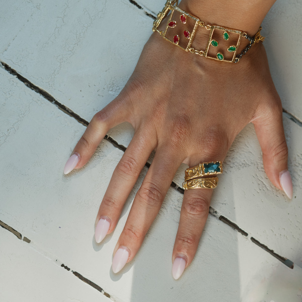 Hand on white painted wood wearing the open square gold link bracelet, ruby red gemstones in one link and emerald green in the next