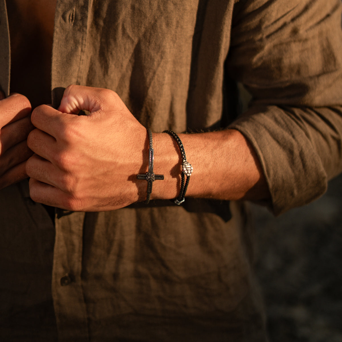 Snake head cord bracelet with ruby eyes worn on a man's wrist in warm evening light beside a black zircon cross cuff