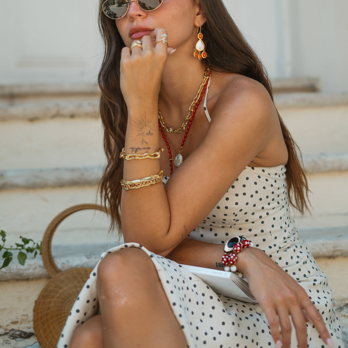 Model seated on stone steps wearing Ebru Jewelry gold bracelets, rings and a coral drop earring