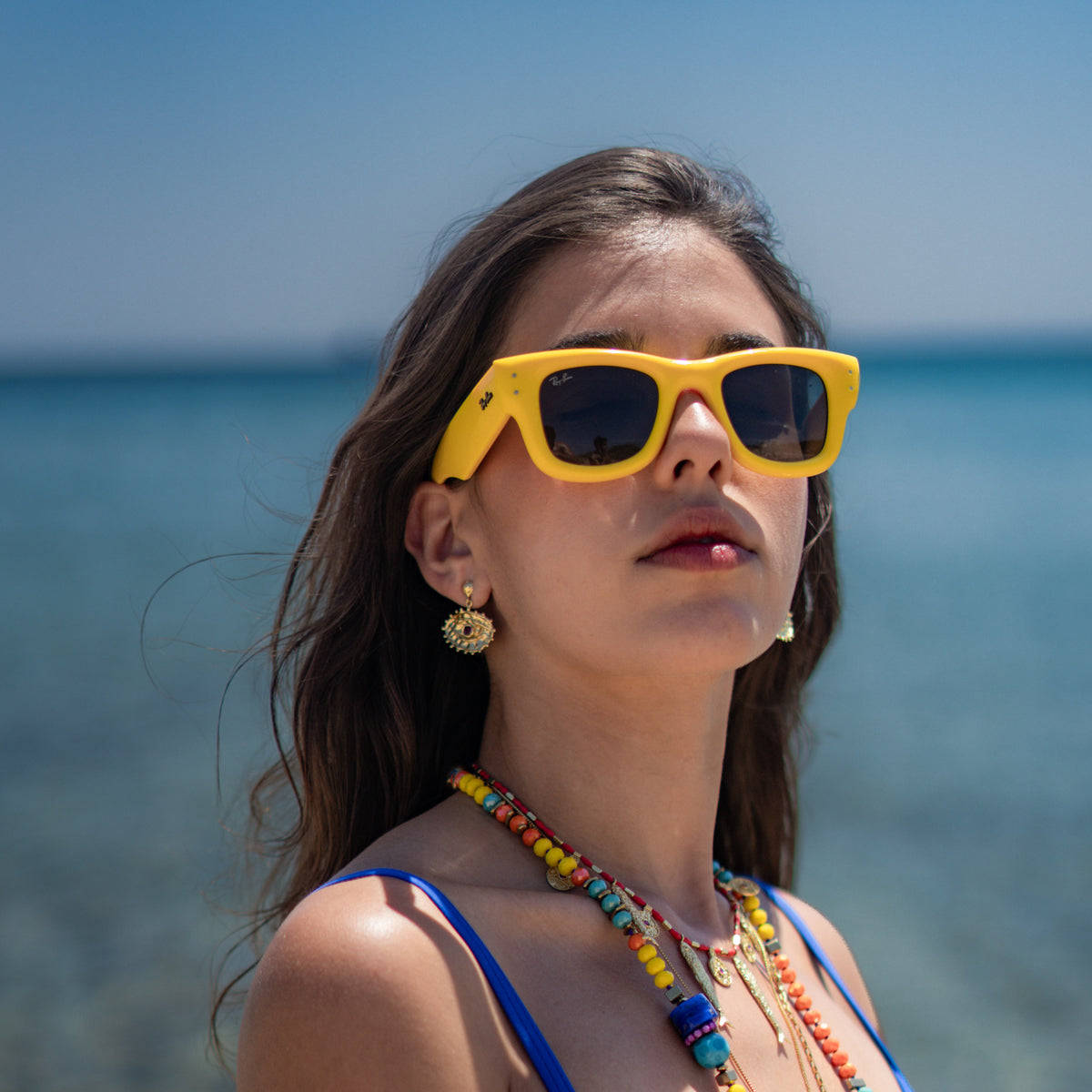 Model looking up toward the sun by the water, the red evil eye gold drop earring catching the light