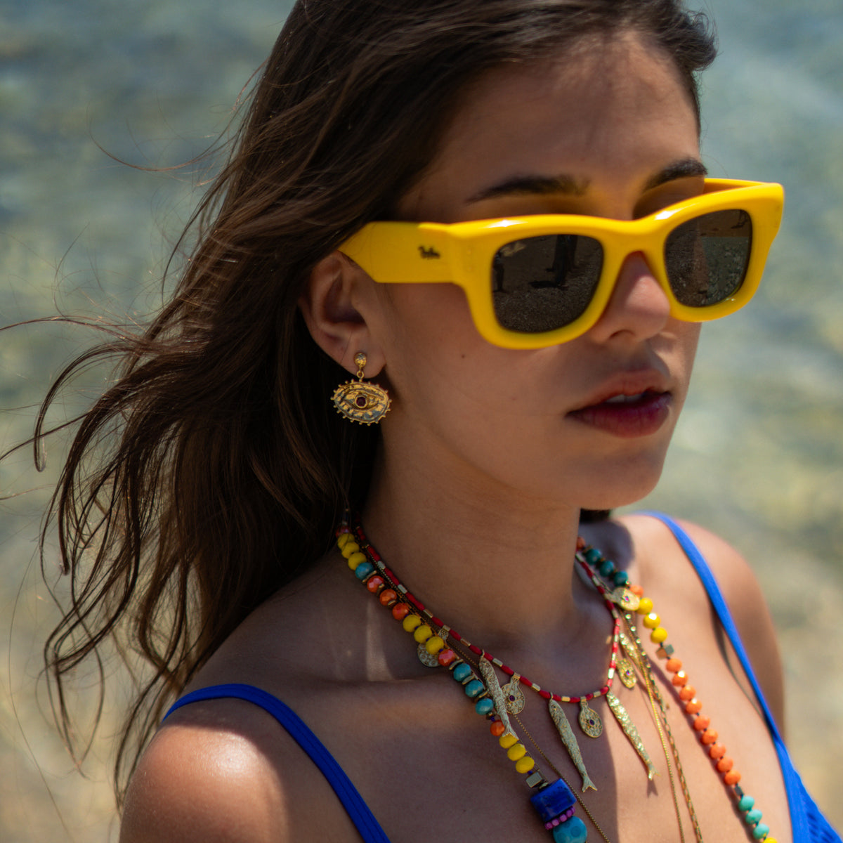 Model by the sea in yellow sunglasses wearing the Sacred Gaze ruby evil eye earring with layered colorful beaded necklaces