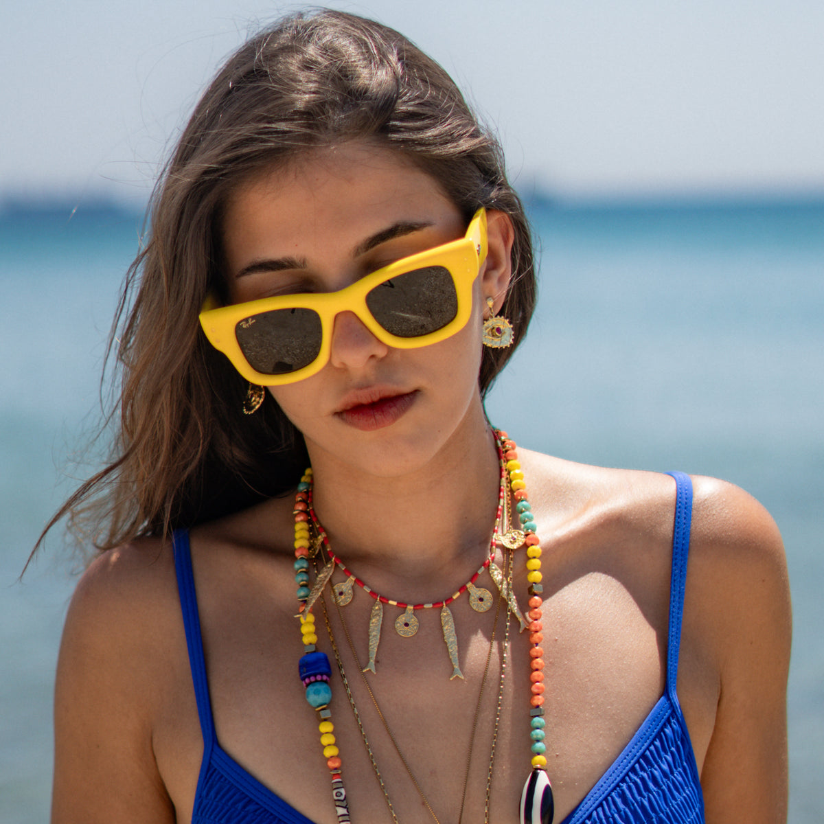 Model with her head tilted on a sunny beach wearing the ruby evil eye gold earrings and colorful layered necklaces