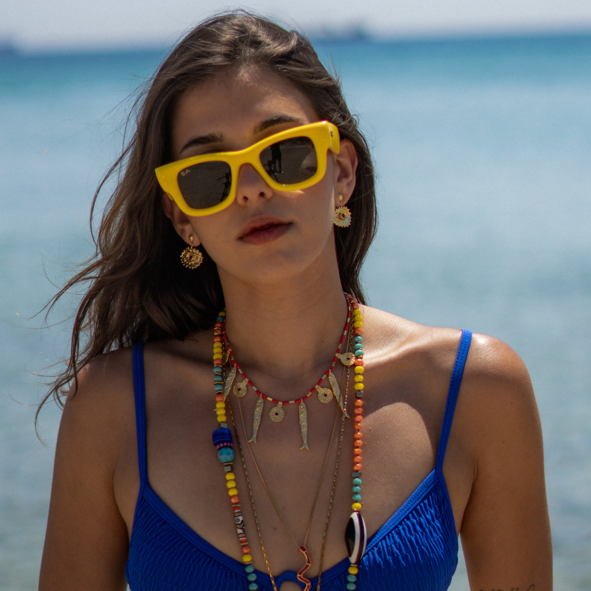 Model in a blue swimsuit on the beach wearing the gold evil eye drop earrings on both ears with stacked beaded and gold fish charm necklaces