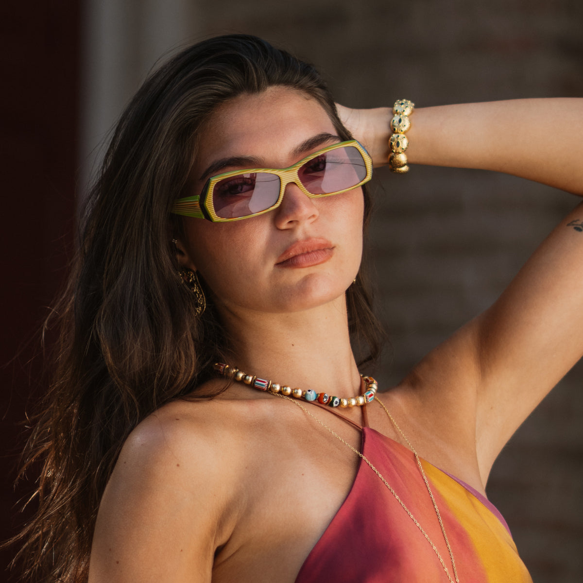 Model in sunglasses and a halter dress with her arm raised, showing the Sapphire Watchful Eye gold cuff bracelet at the wrist