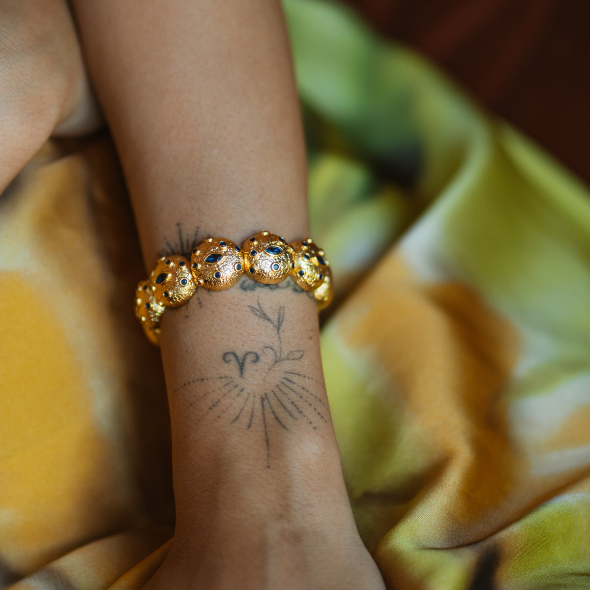 Textured dome gold cuff bracelet with blue zircon eyes worn on a tattooed wrist against yellow and green silk