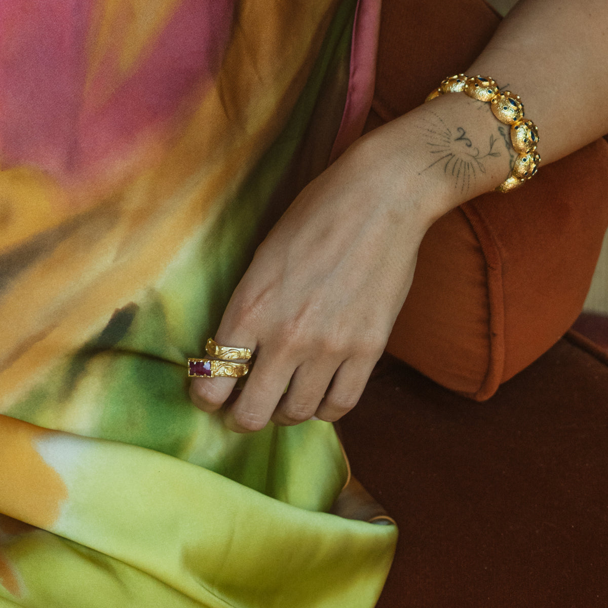 Gold textured dome cuff bracelet on a wrist resting on a rust velvet chair, with stacked gold rings on the hand