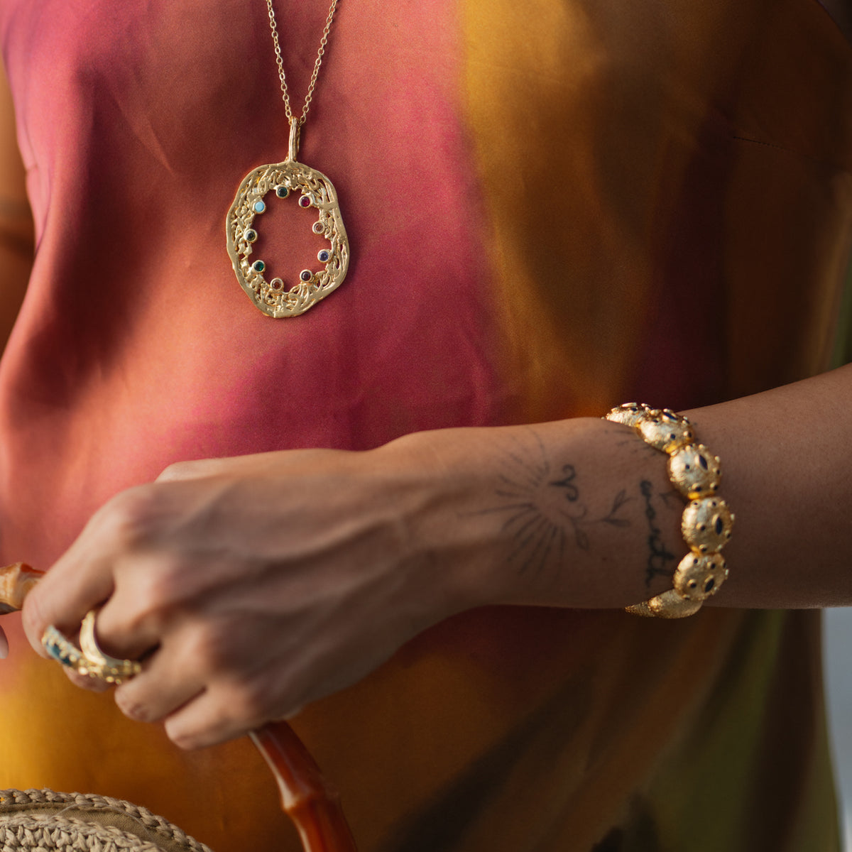 Close view of the gold dome cuff bracelet on a tattooed wrist beside a gold oval pendant with multicolor gemstones and a woven straw bag