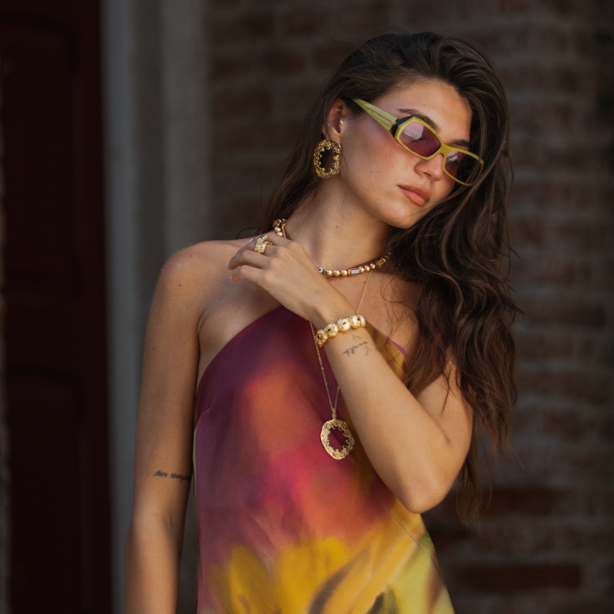 Model in a pink and yellow halter dress wearing the Watchful Eye gold cuff bracelet with a gold oval gemstone pendant necklace