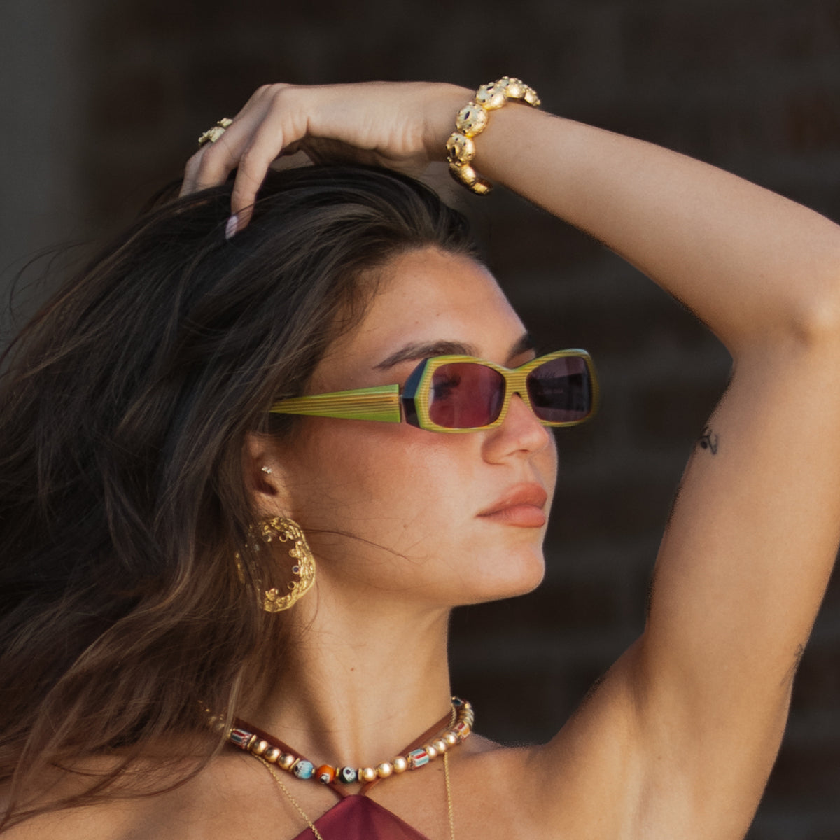 Model in yellow sunglasses with one hand in her hair, wearing the gold dome cuff bracelet, filigree oval earrings and a beaded choker