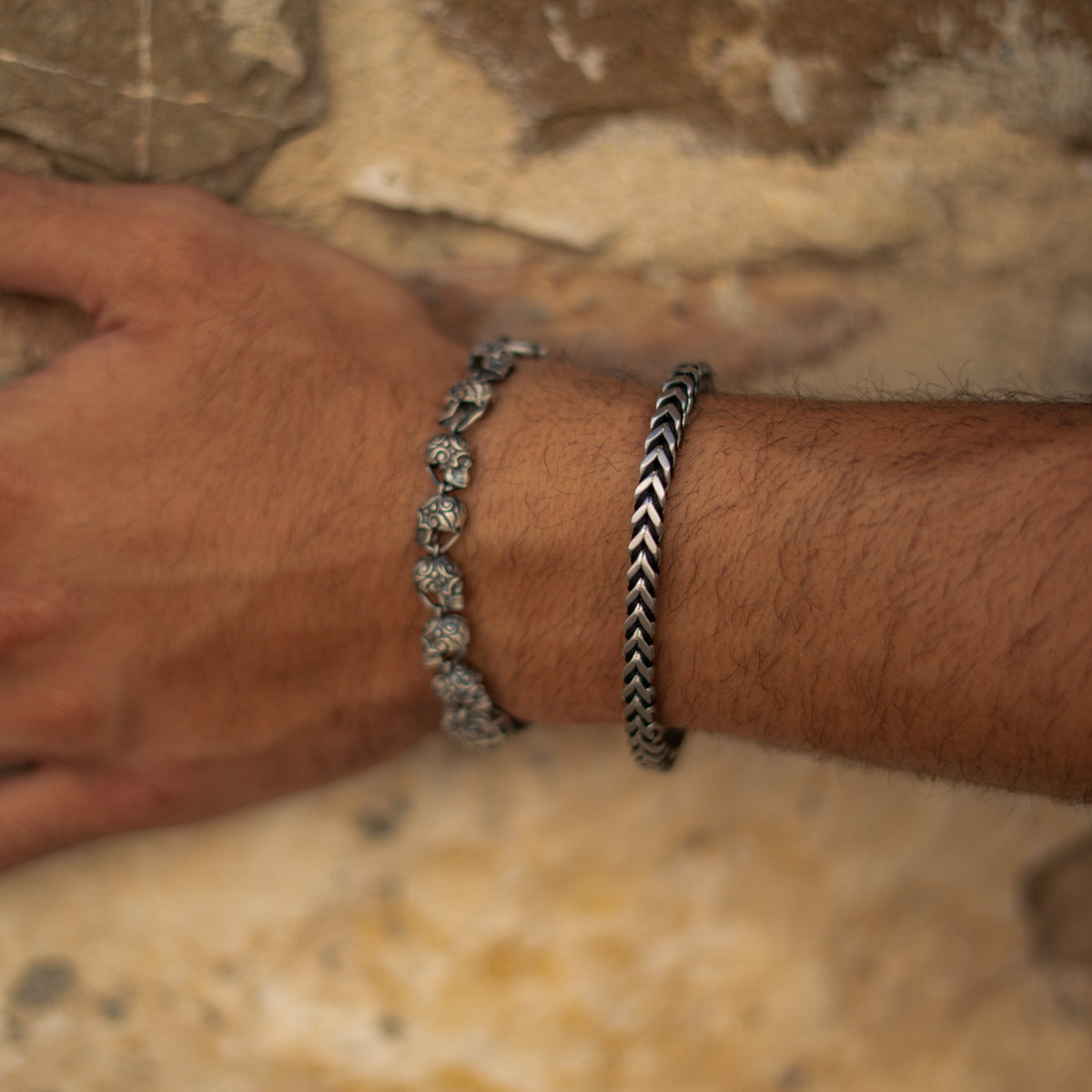 Men's skull link sterling silver bracelet worn beside a chevron chain bracelet against a stone wall