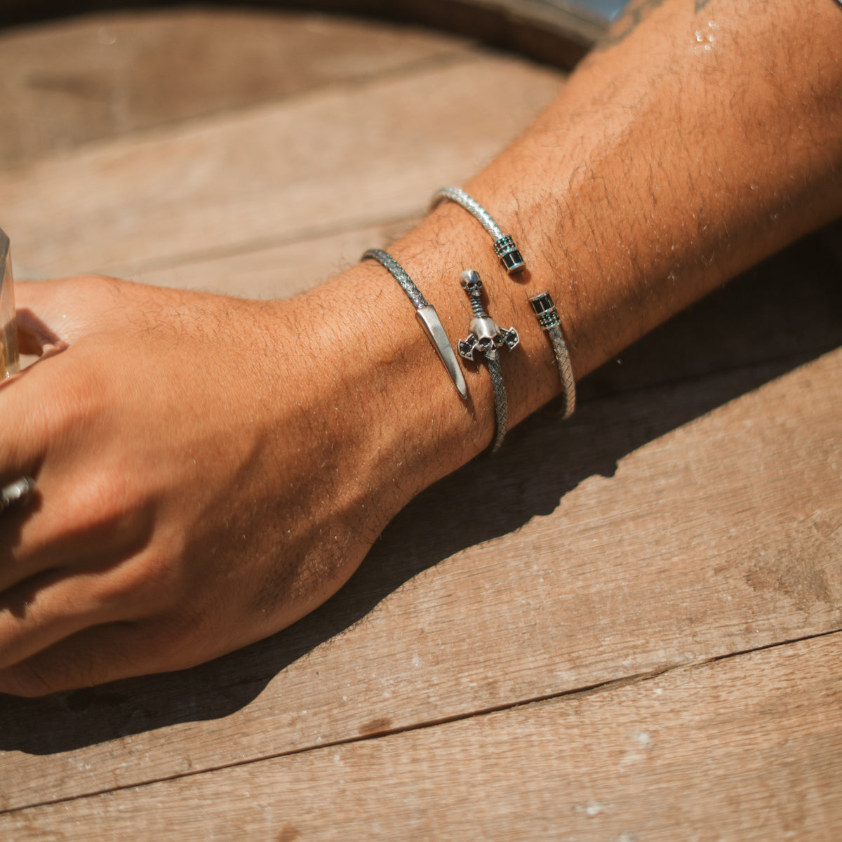 Man's arm resting on a wooden table in sunlight, wearing the skull and dagger oxidized silver wrap bracelet