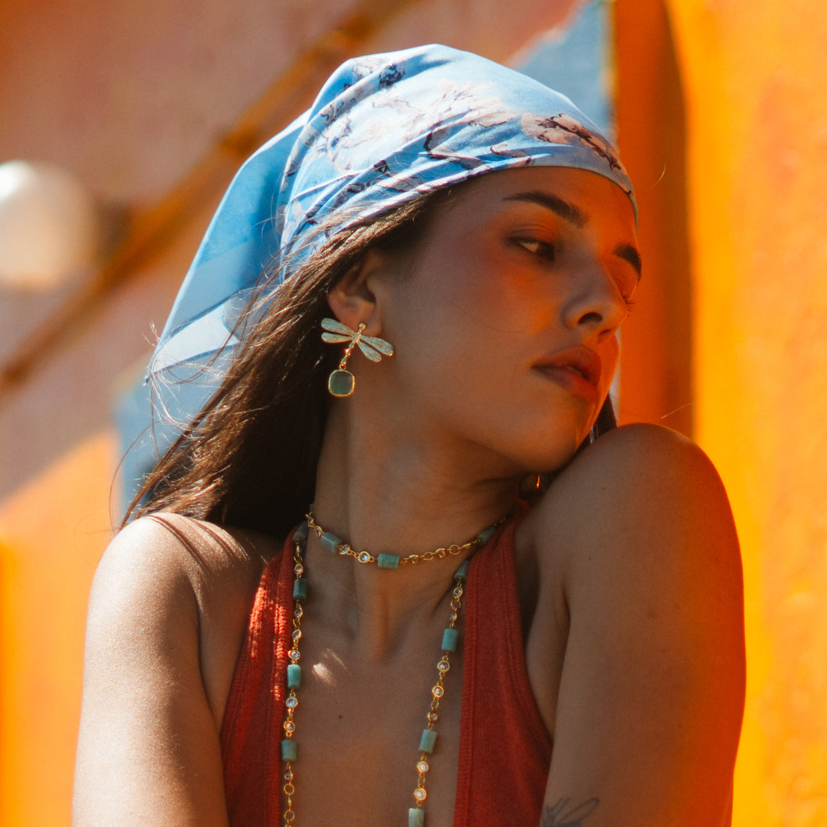 Model in a blue silk headscarf and rust top against an orange wall, wearing the gold dragonfly aqua cat's eye earring and beaded necklaces